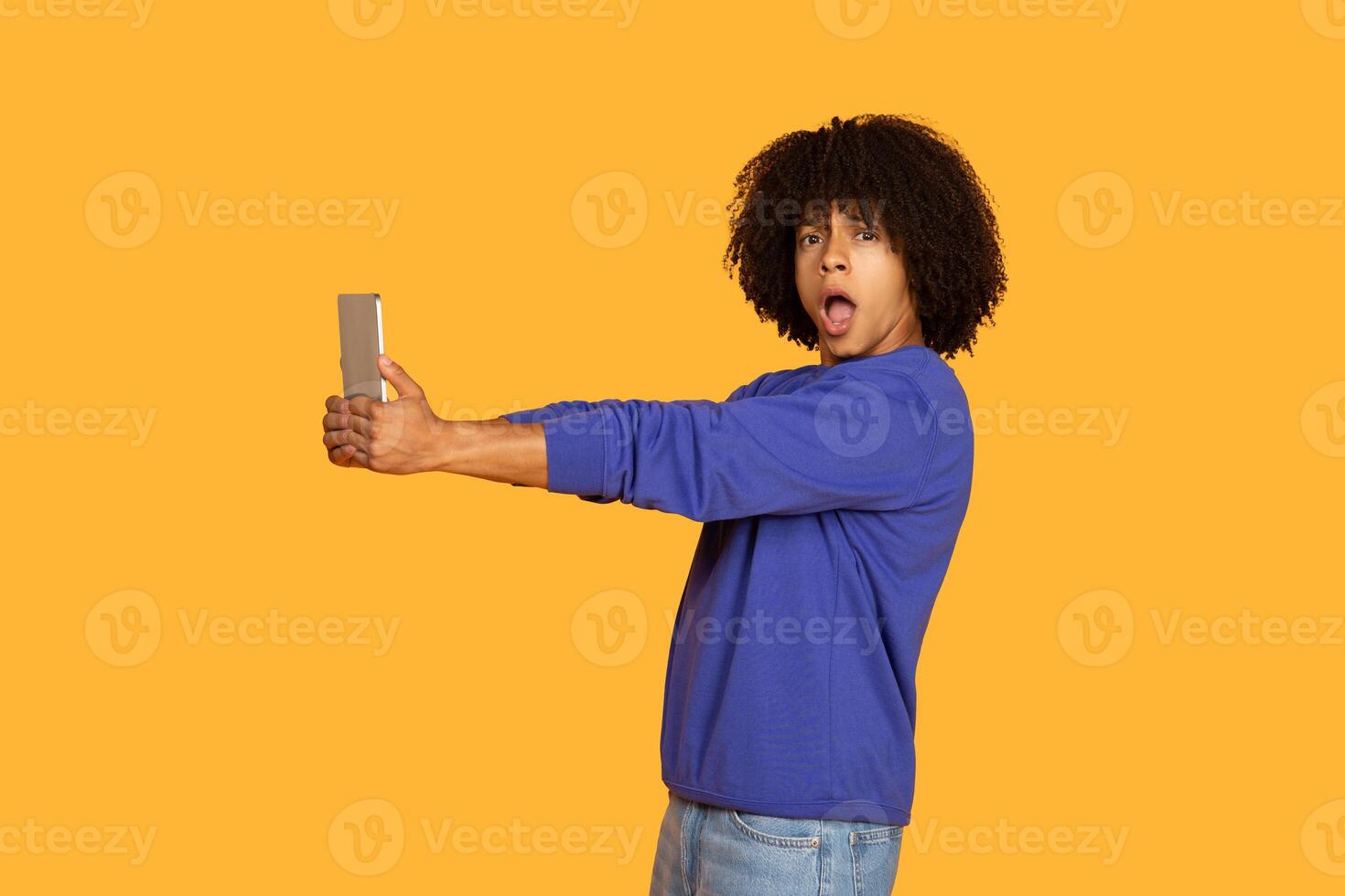 A young man with curly hair is taking a selfie with tablet, looking shocked and surprised, wearing a blue sweater and jeans, standing against a bright yellow background. photo