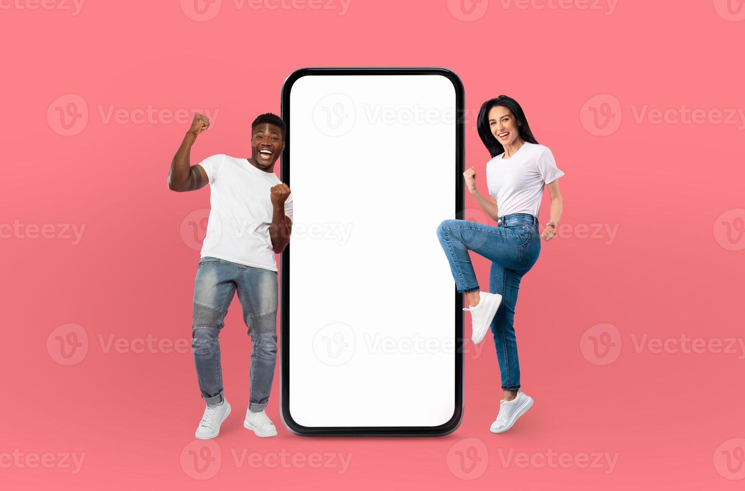 A smiling multiethnic young man and woman stand confidently beside a large smartphone with a blank screen, isolated on a vivid blue background. photo