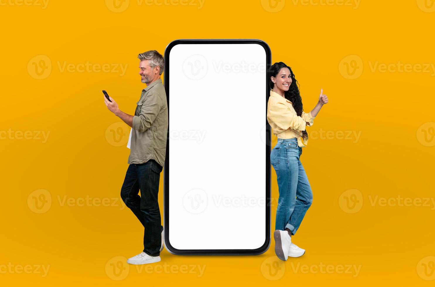Cheerful European couple leaning on huge smartphone with white blank screen, posing isolated on yellow studio background, mockup copy space photo