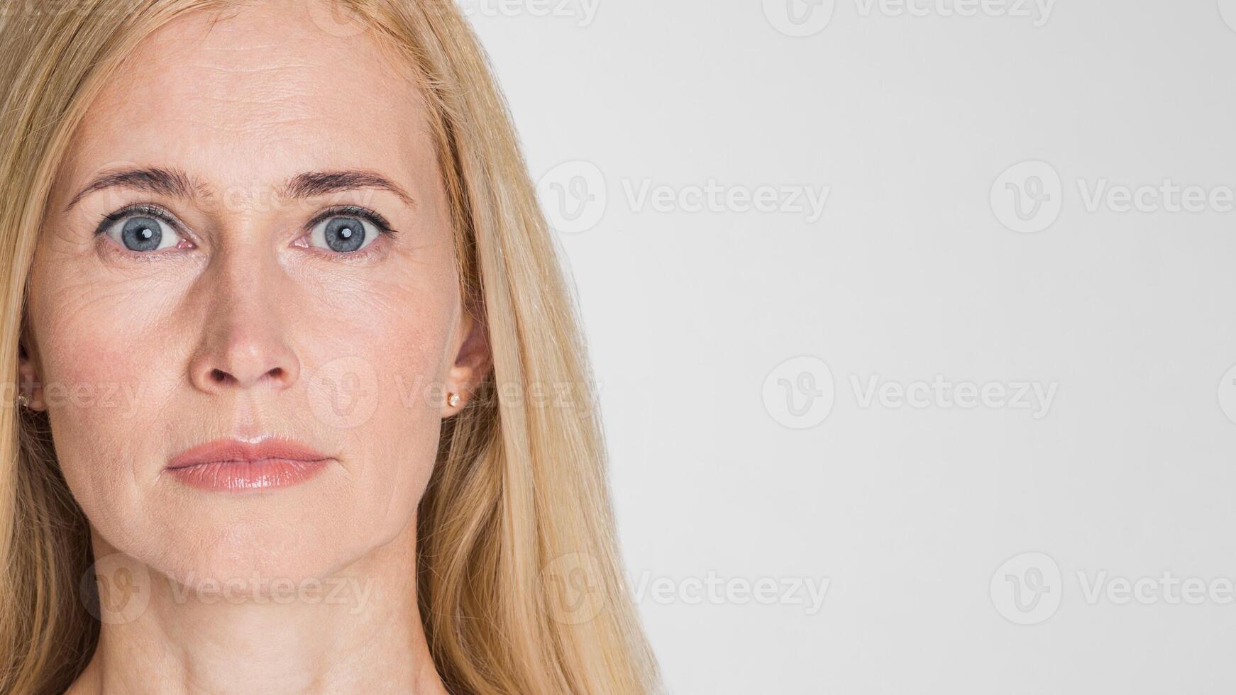 This is a close-up portrait of a blonde woman with blue eyes looking directly at the camera. She has a serious expression on her face, with her lips slightly pursed. The background is plain white photo