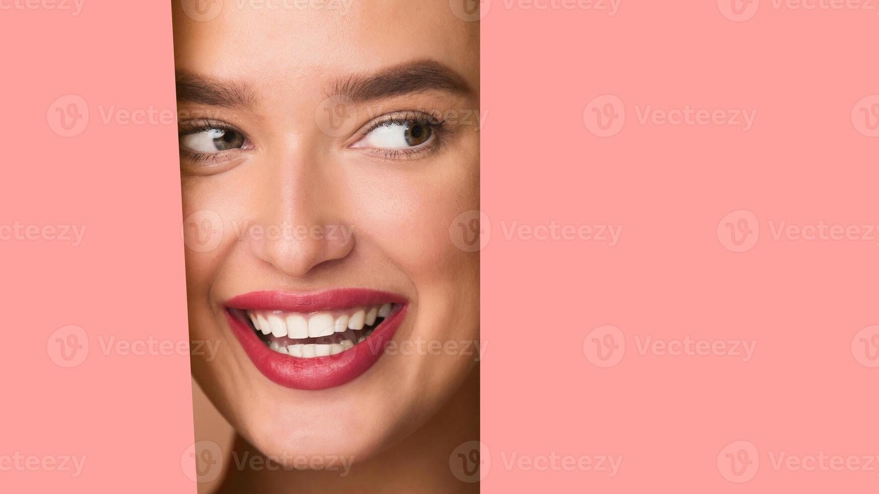 A woman with a joyful expression is smiling through a hole in a vibrant pink background. Her face is visible, displaying happiness and delight in an unexpected setting, empty space photo