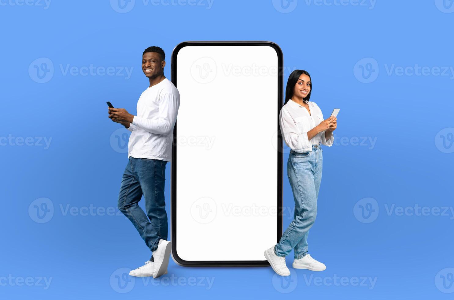 On a deep blue background, a multiracial couple, both smiling, stands adjacent to an enormous smartphone with a blank display, ready for showcasing apps or designs. photo