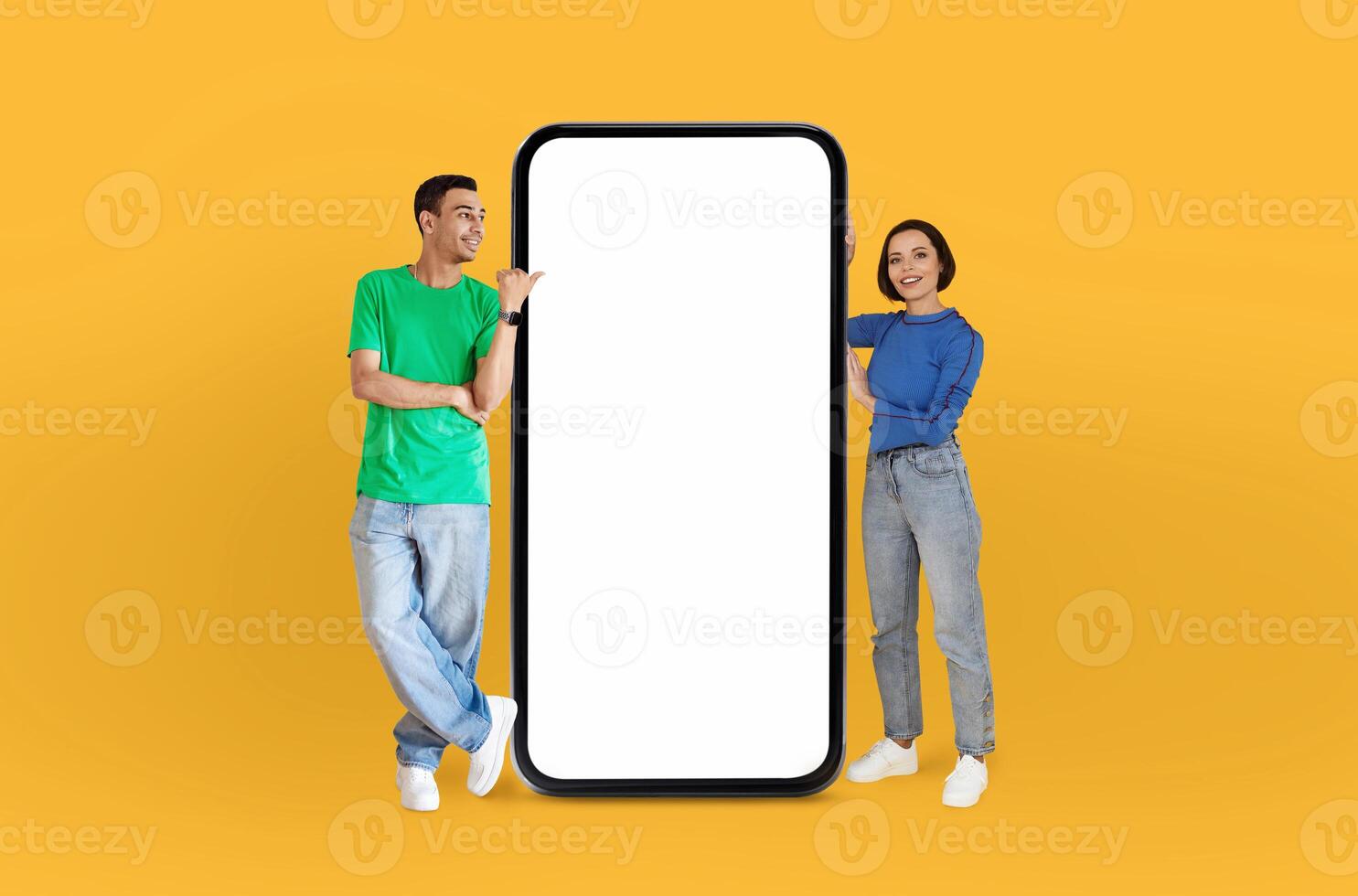 Energetically, the young multiethnic couple engages with viewers, standing next to a mockup smartphone that offers a clear, unoccupied screen for various displays. photo
