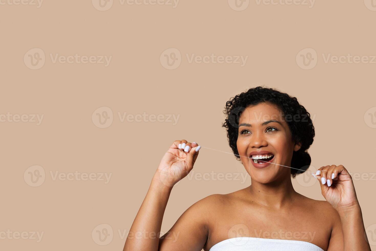 A cheerful woman with short, curly hair is captured mid-floss, taking care of her dental hygiene. She is wrapped in a towel, suggesting a setting possibly post-shower, copy space photo