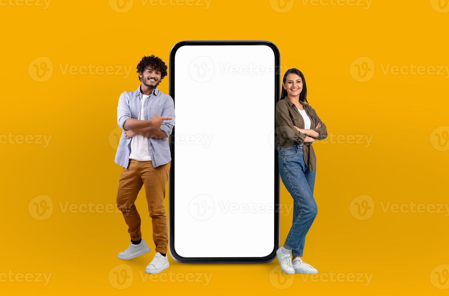 A cheerful young hindu man and woman stand confidently next to a large, blank screen smartphone that dominates the center of the frame, suggesting a technology theme photo