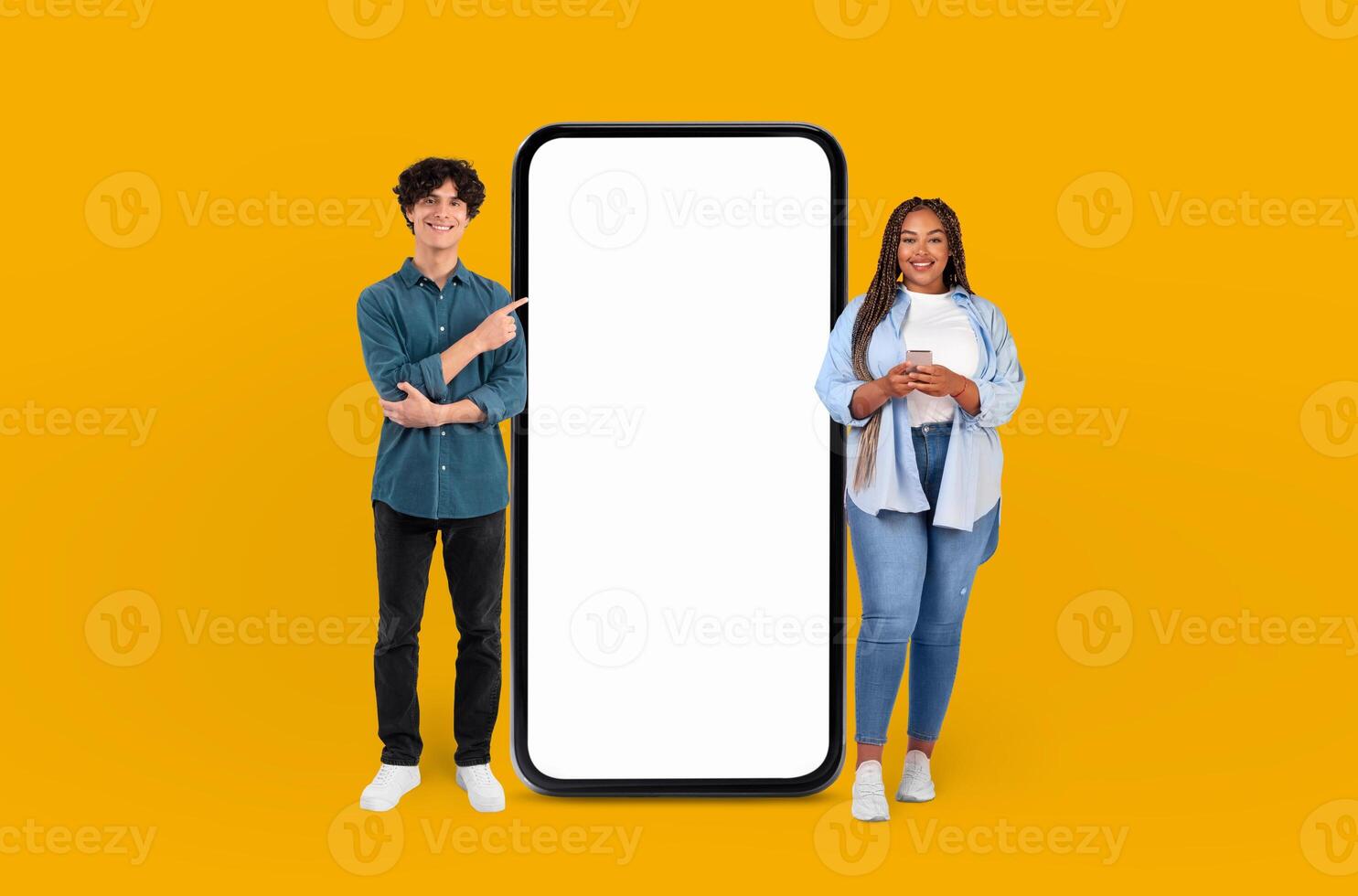 The vibrant yellow background highlights the smiling multiracial young couple as they gesture towards the expansive blank screen of the smartphone beside them. photo