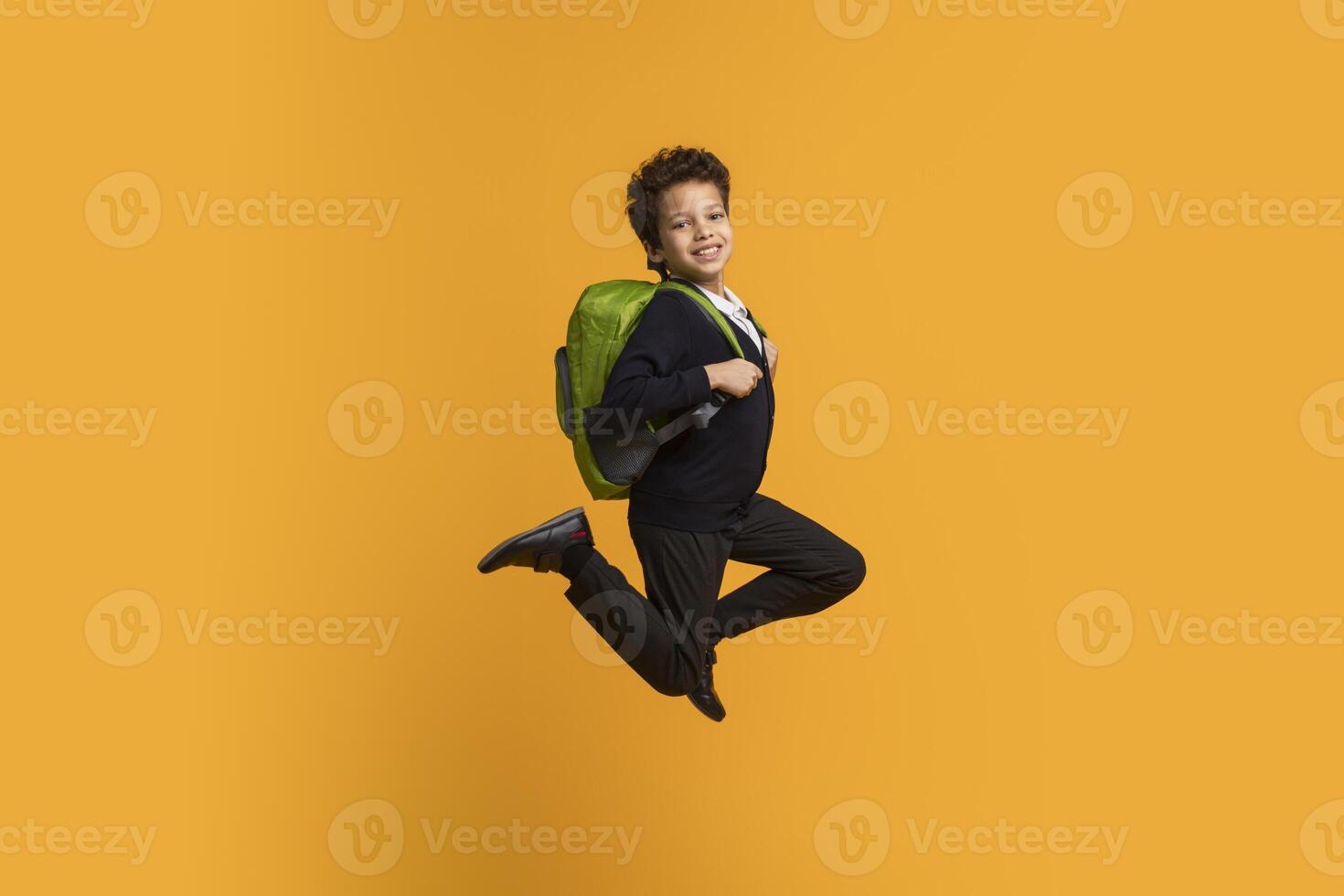 African American boy wearing a black blazer and black pants is jumping in front of a bright yellow background. He has a green backpack on and is smiling at the camera. photo