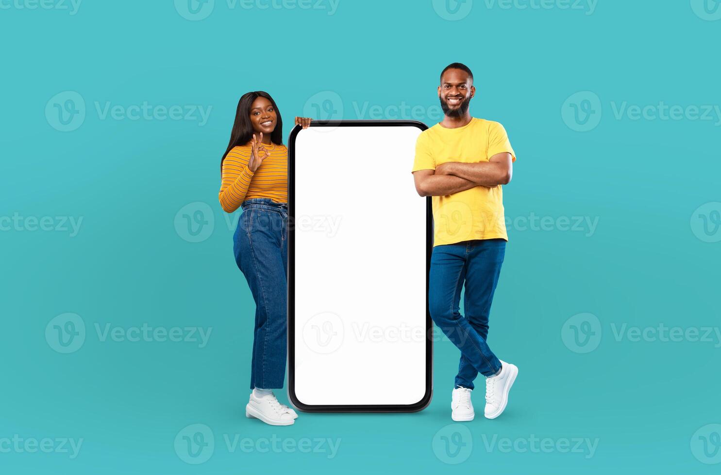 A young black man and woman are standing next to a blank screen against a blue background. They are posing confidently, looking directly at the camera. photo