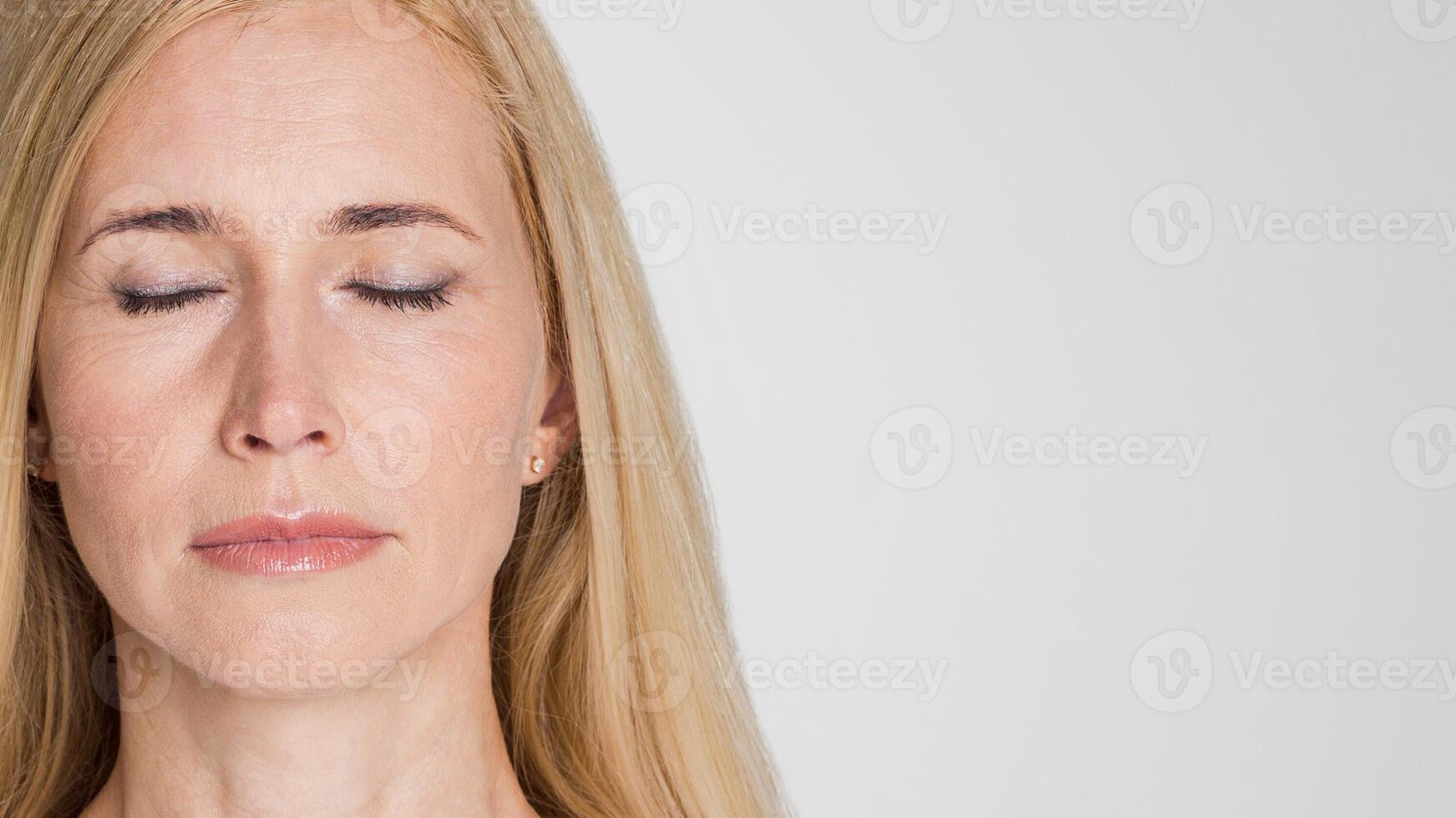 A close-up portrait of a woman with blonde hair, wearing a simple white shirt, looking at the camera with her eyes closed. The image captures her soft features and calm expression, copy space photo