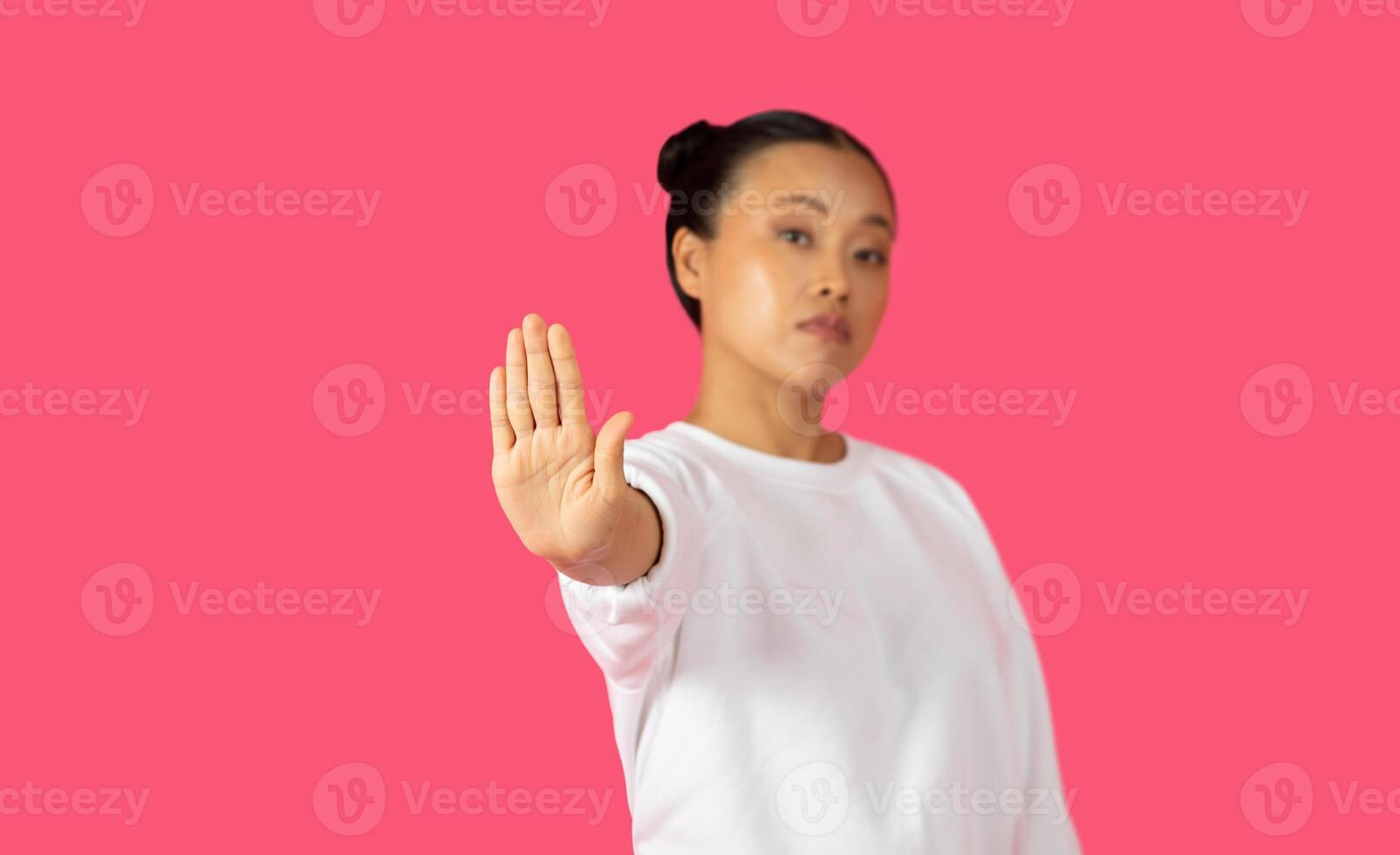 Asian young woman with black hair tied up in a bun is wearing a white sweatshirt and standing in front of a pink background. Her hand is held up in a stop gesture, with her palm facing the camera. photo