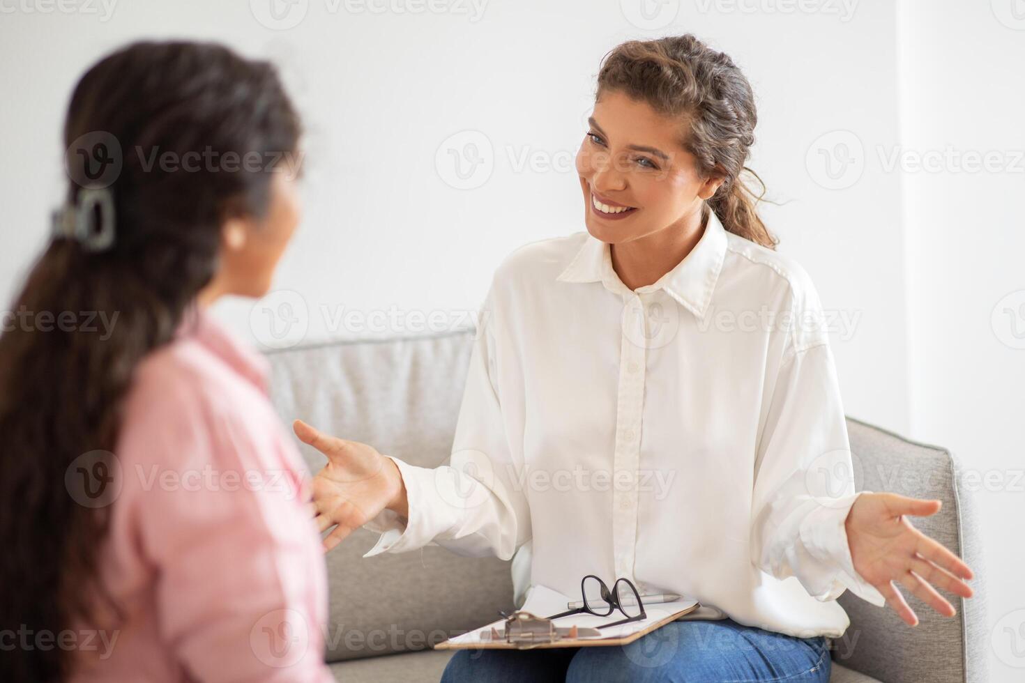 A woman in a white shirt is sitting on a couch and talking to a client. She is gesturing with her hands and smiling. The client is listening intently. This image depicts a typical therapy session photo