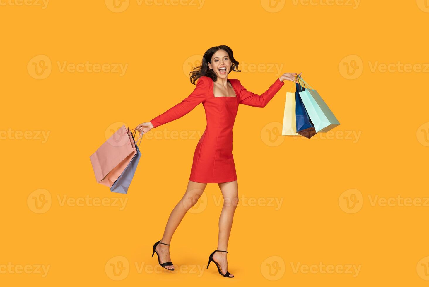 A happy woman in a red dress walks on a yellow background while carrying multiple shopping bags in her hands. She smiles at the camera and seems excited about her purchases. photo