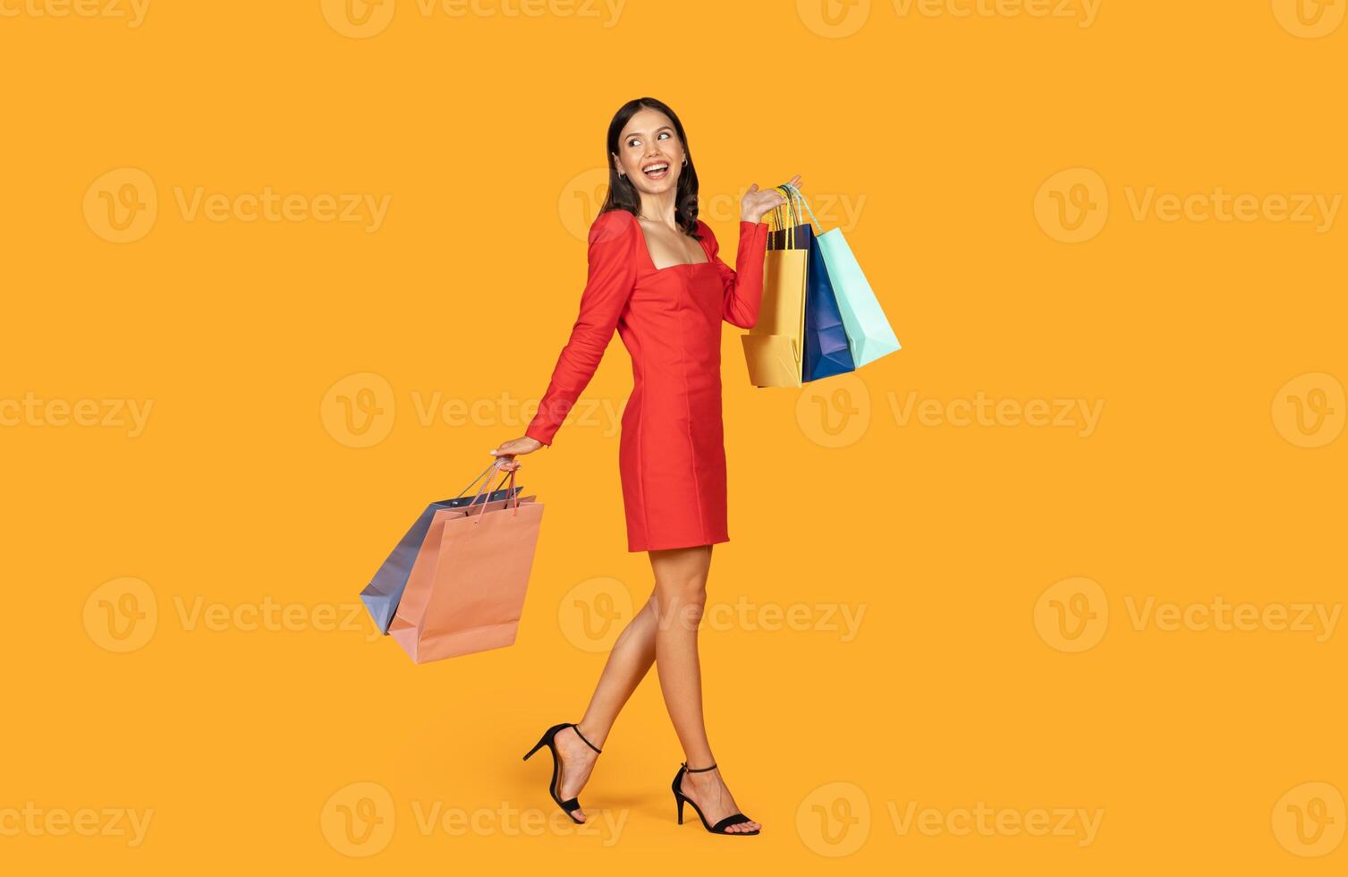 A woman wearing a red dress walks with shopping bags in her hands in front of a yellow background. She is smiling and looks happy with her purchases. photo