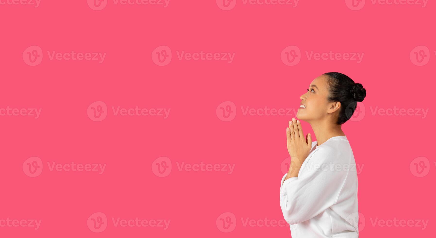 Asian woman with her hands clasped together in prayer looks upward with a hopeful expression. Her dark hair is pulled back in a bun, and she is wearing a white blouse, copy space photo