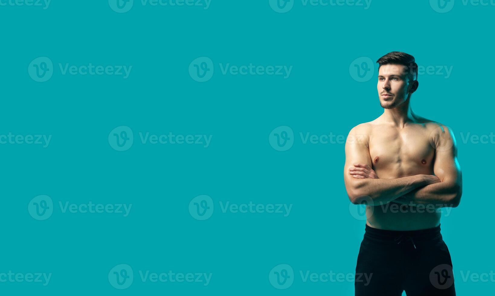 A shirtless man is standing confidently in front of a vibrant turquoise background. His muscular build is prominent as he poses with his arms at his sides, copy space photo
