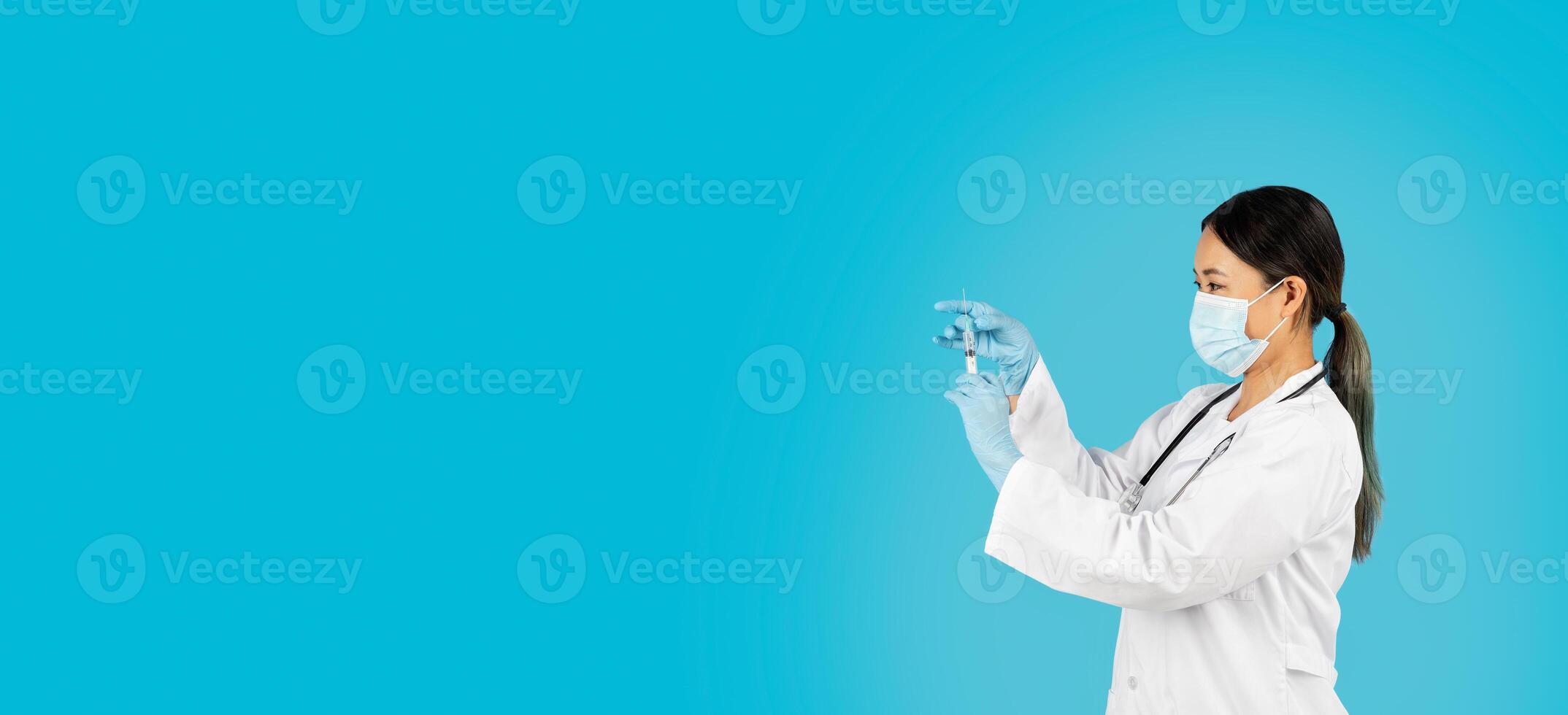 Asian woman doctor, wearing a white coat, a blue face mask, and blue gloves, is holding a syringe in her right hand. She is looking at the syringe. The background is a solid blue color, copy space photo