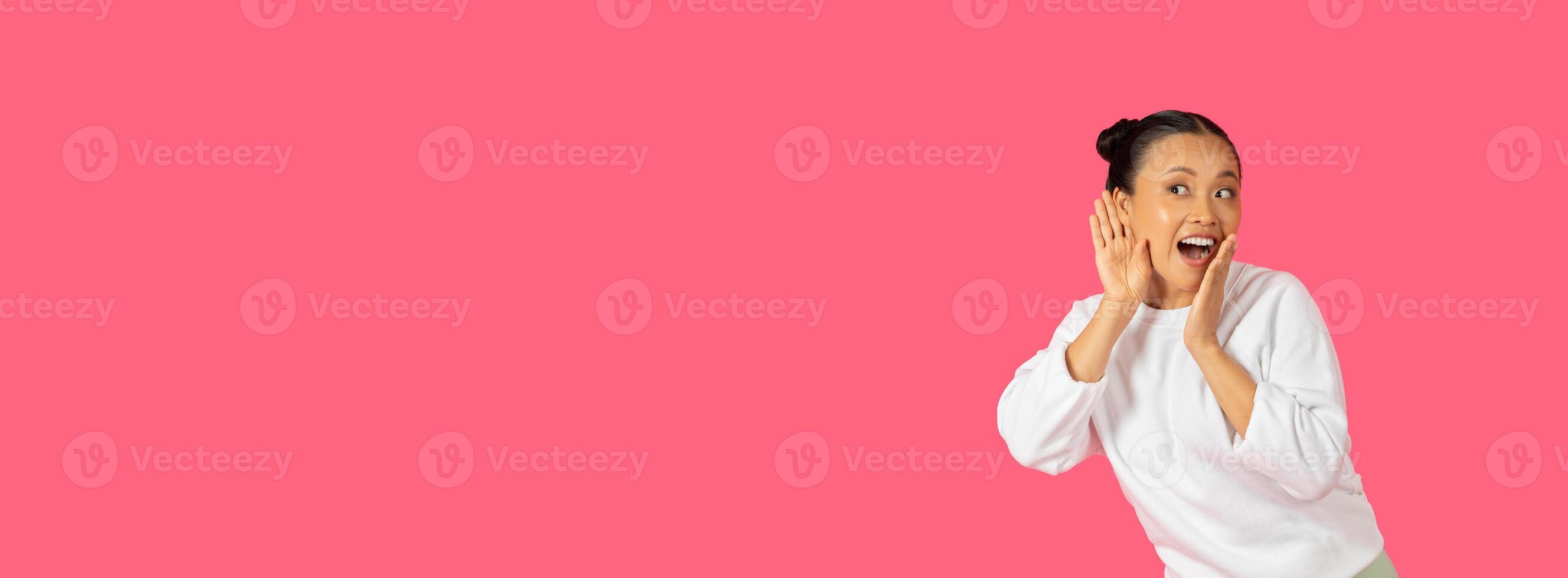 A joyful Asian lady stands against a vibrant pink backdrop, enthusiastically gesturing as if sharing exciting news, panorama with copy space photo