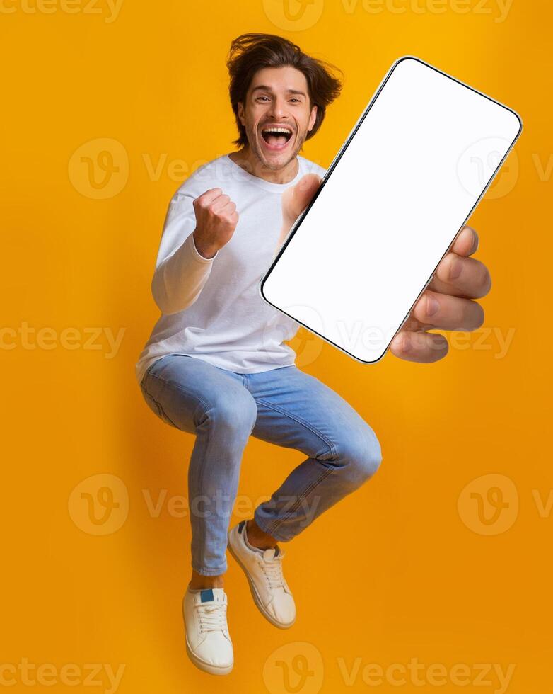 New awesome mobile app. Jumping emotional young guy showing smartphone with blank screen on orange studio background, mockup for application or website design photo