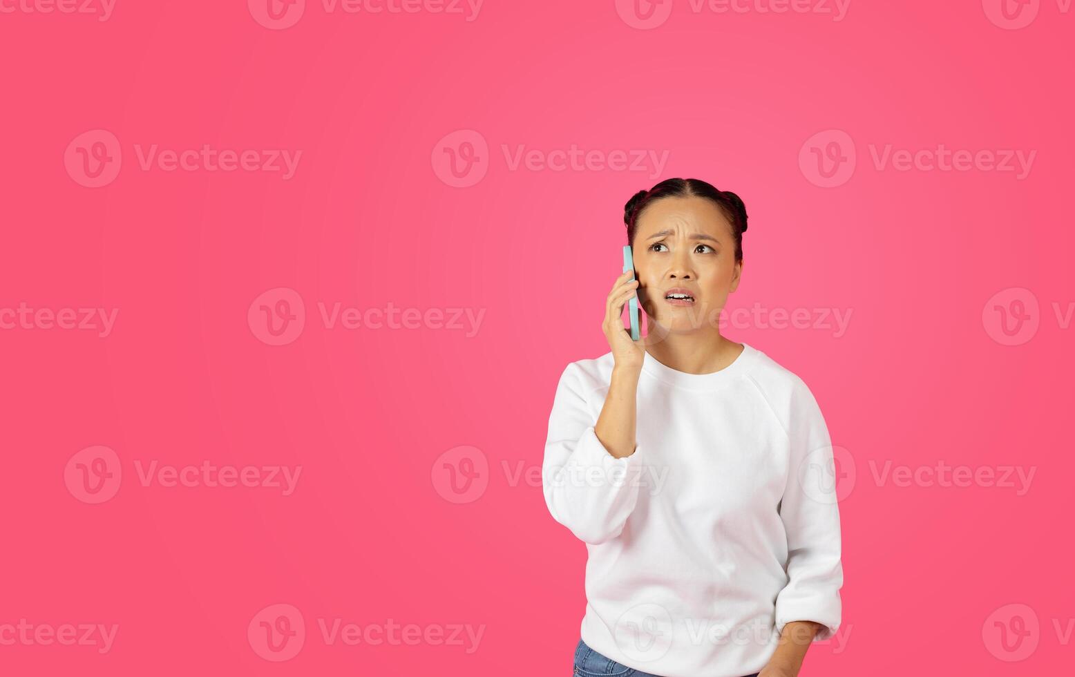 Asian woman with her hair in buns is talking on a cell phone, and she has a confused expression on her face. The background is a solid pink color, copy space photo