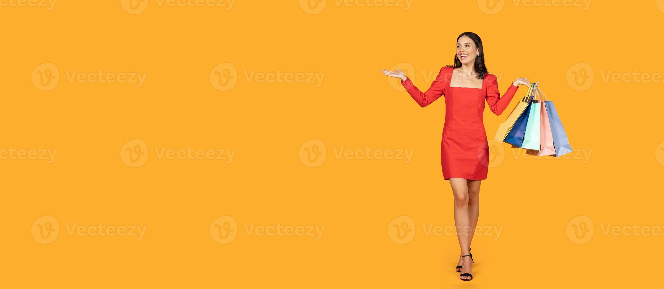 A woman in a red dress is standing in front of a yellow background. She is holding a stack of colorful shopping bags in her right hand and gesturing with her left hand, panorama with copy space photo