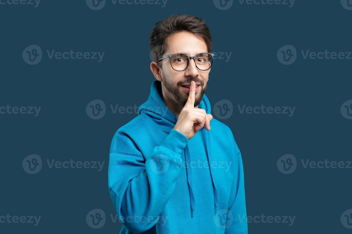 A man in a bright blue hoodie stands against a matching blue background, placing one finger over his lips as if to gesture for silence or secrecy. photo