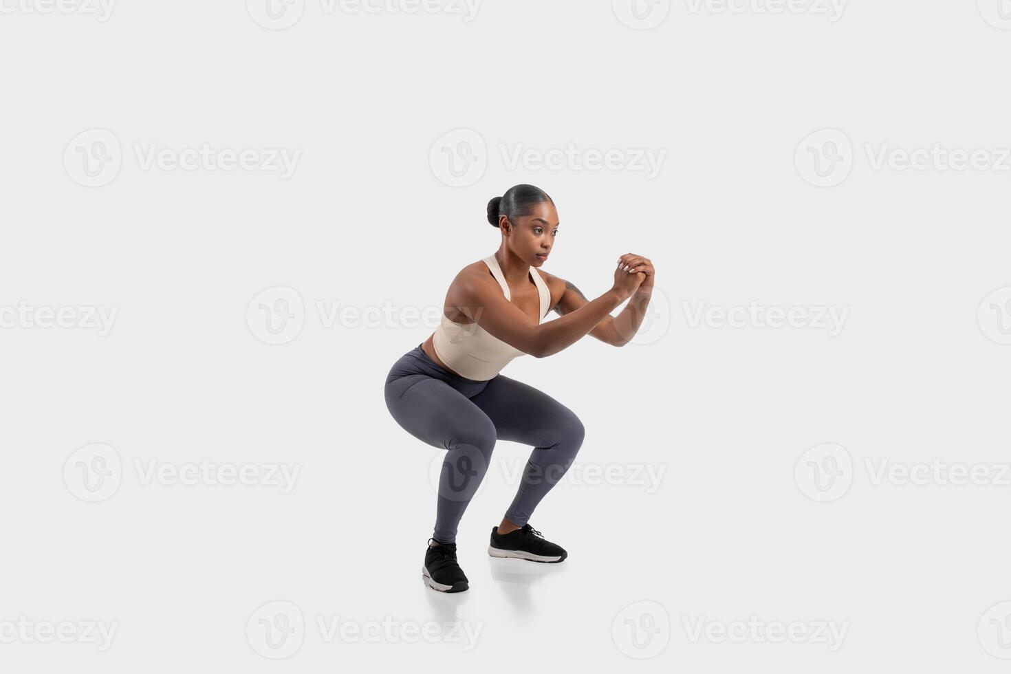 Black woman with dark hair pulled back in a bun wears a white tank top and grey leggings as she performs a squat exercise. Her fists are clenched at her chest and she is looking straight ahead photo