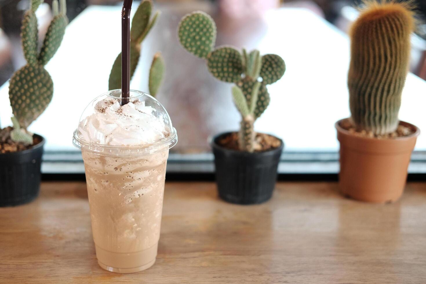 Fresh ice latte frappe coffee with bulb in plastic glass on wooden table and cactus plant decorate on counter in cafe. Copy space photo