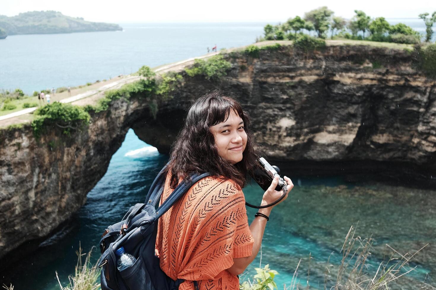 Smiling Asian woman is holding mirrorless camera and rocky cliff at the sea for happiness and destination travel on Broken Beach in Bali island at Indonesia photo