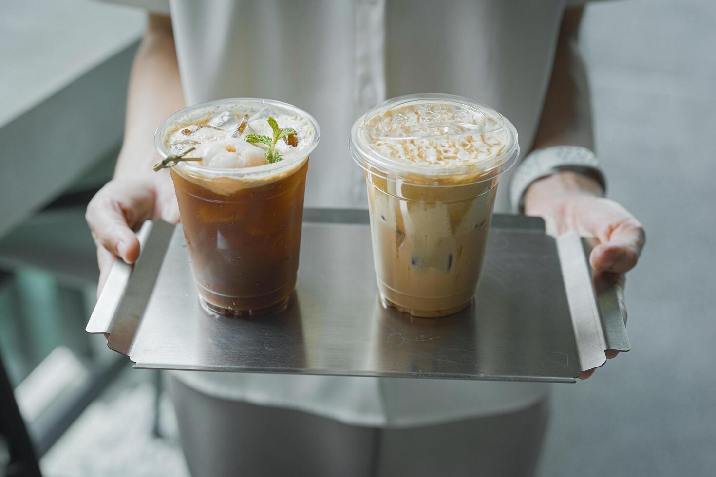 Close up and selective focus hand of employee girl holding tray of ice lychee americano and cappuccino at cafe. photo