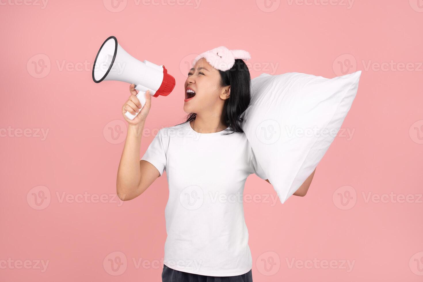 Sad and crazy Asian woman in pajamas and blindfold hugging a pillow. She is screaming into a sound amplifier on pastel pink background in studio. Upset. photo
