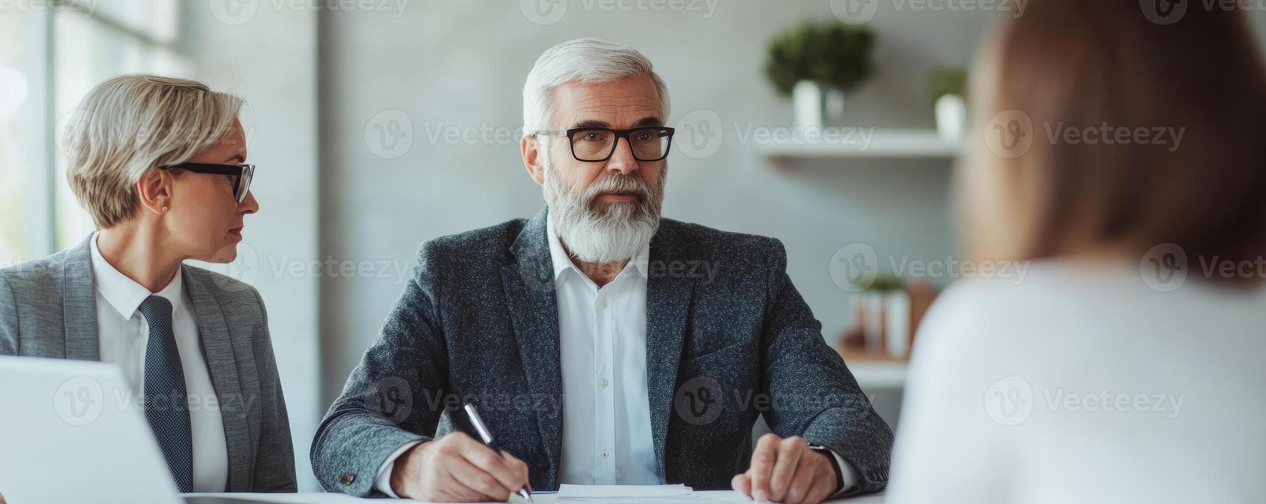 A senior businessman with a beard engages in a professional meeting, flanked by two colleagues, discussing strategies with focused expressions in a modern workspace. photo