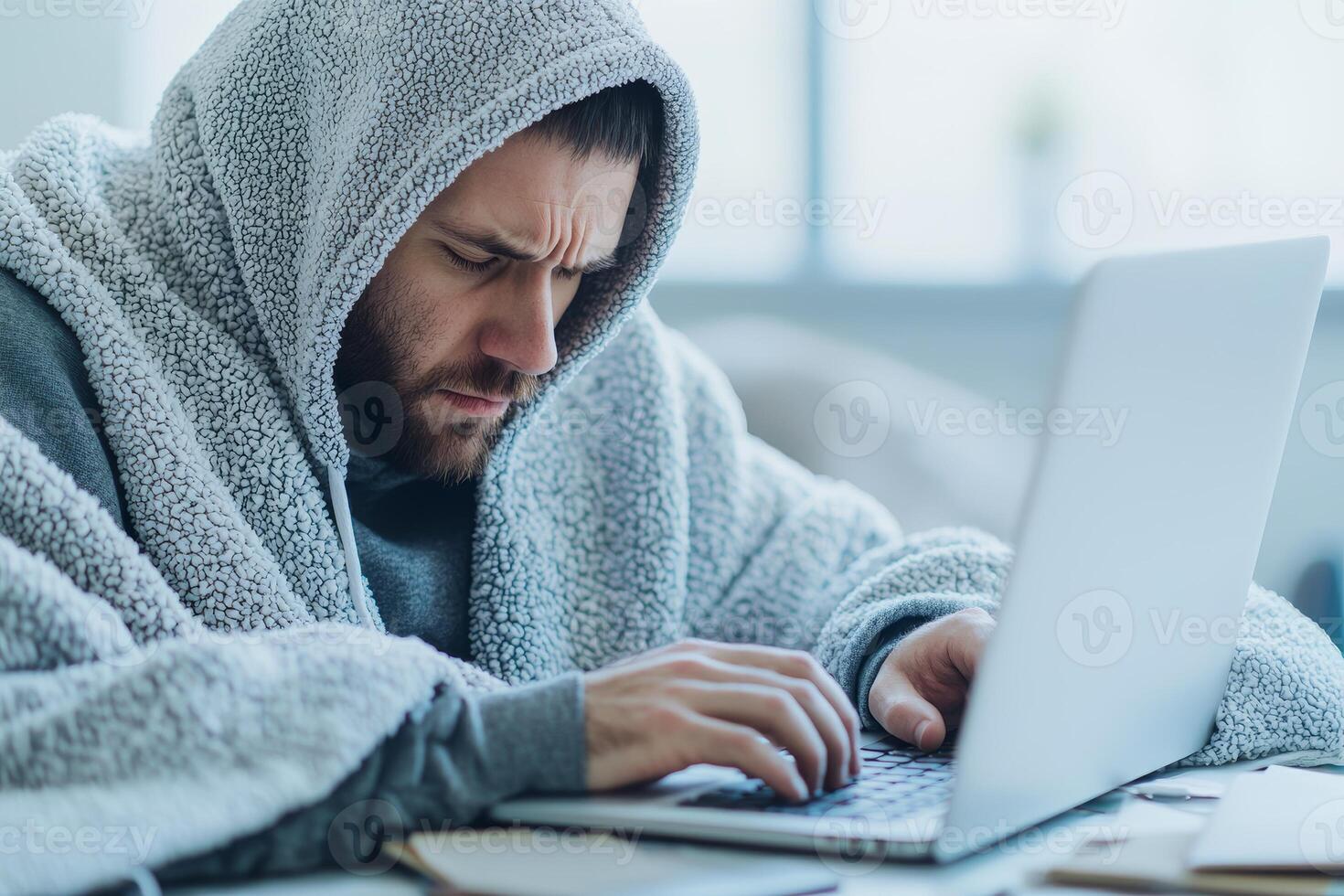 Man under blanket working on laptop with focused expression at home. photo