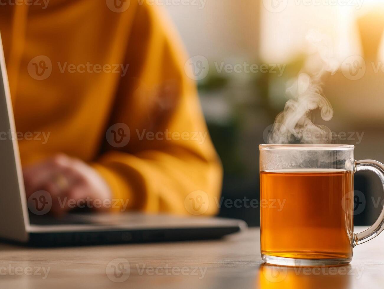 Steaming cup of tea beside a laptop, cozy workspace in warm light. photo