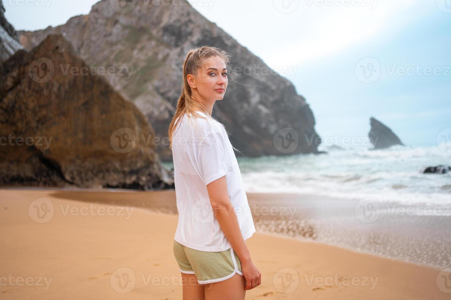 Confident sporty woman standing against huge rock on seashore at beach photo