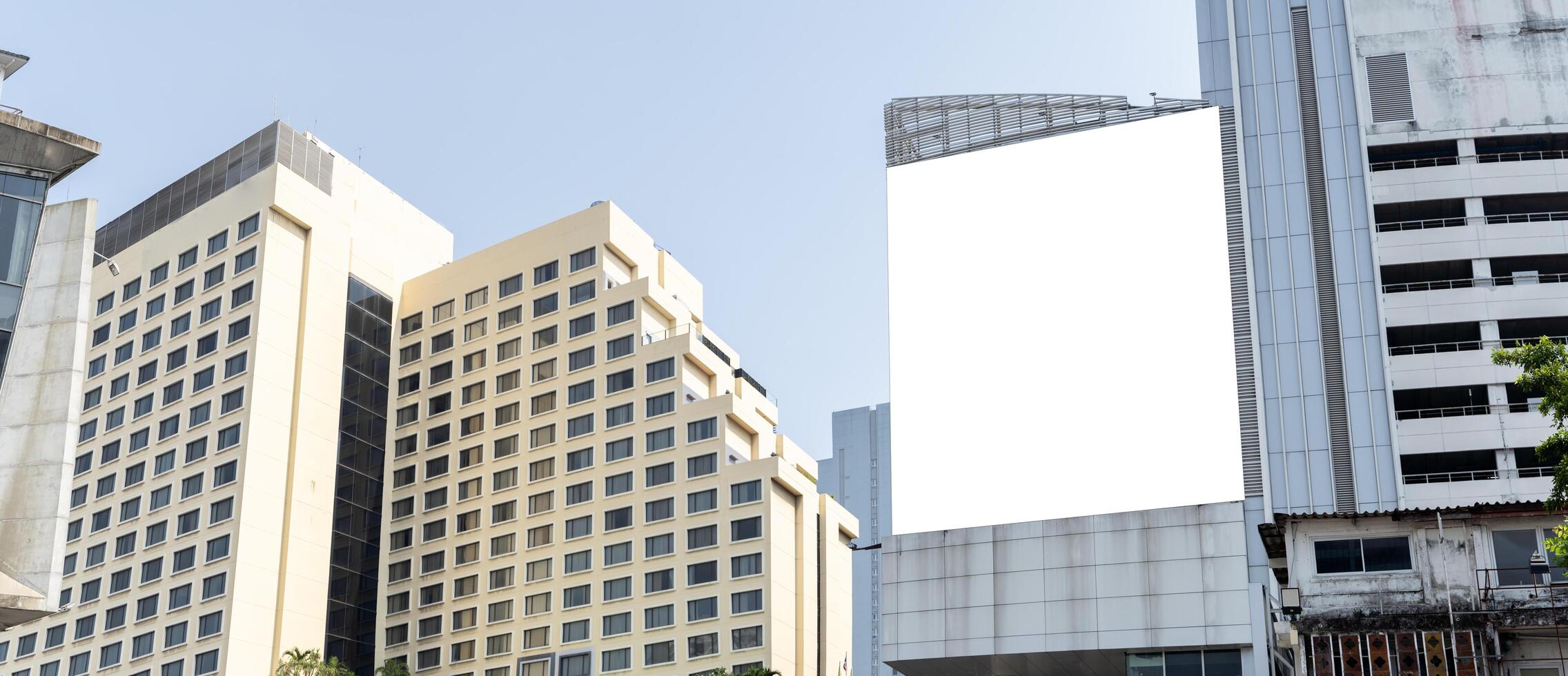 A large blank billboard hangs on the side of a tall building in a city. The billboard is white and has a blue sky in the background. photo