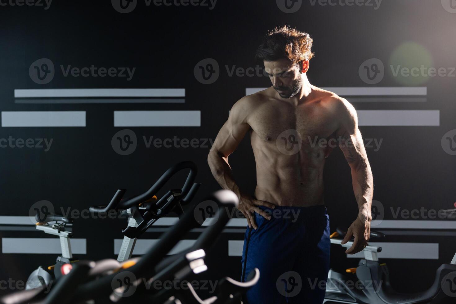 Shirtless caucasian muscular sport man with perfect abs and bicep muscle inside gym with dark background for exercising and workout in high contrast low key lighting studio photo