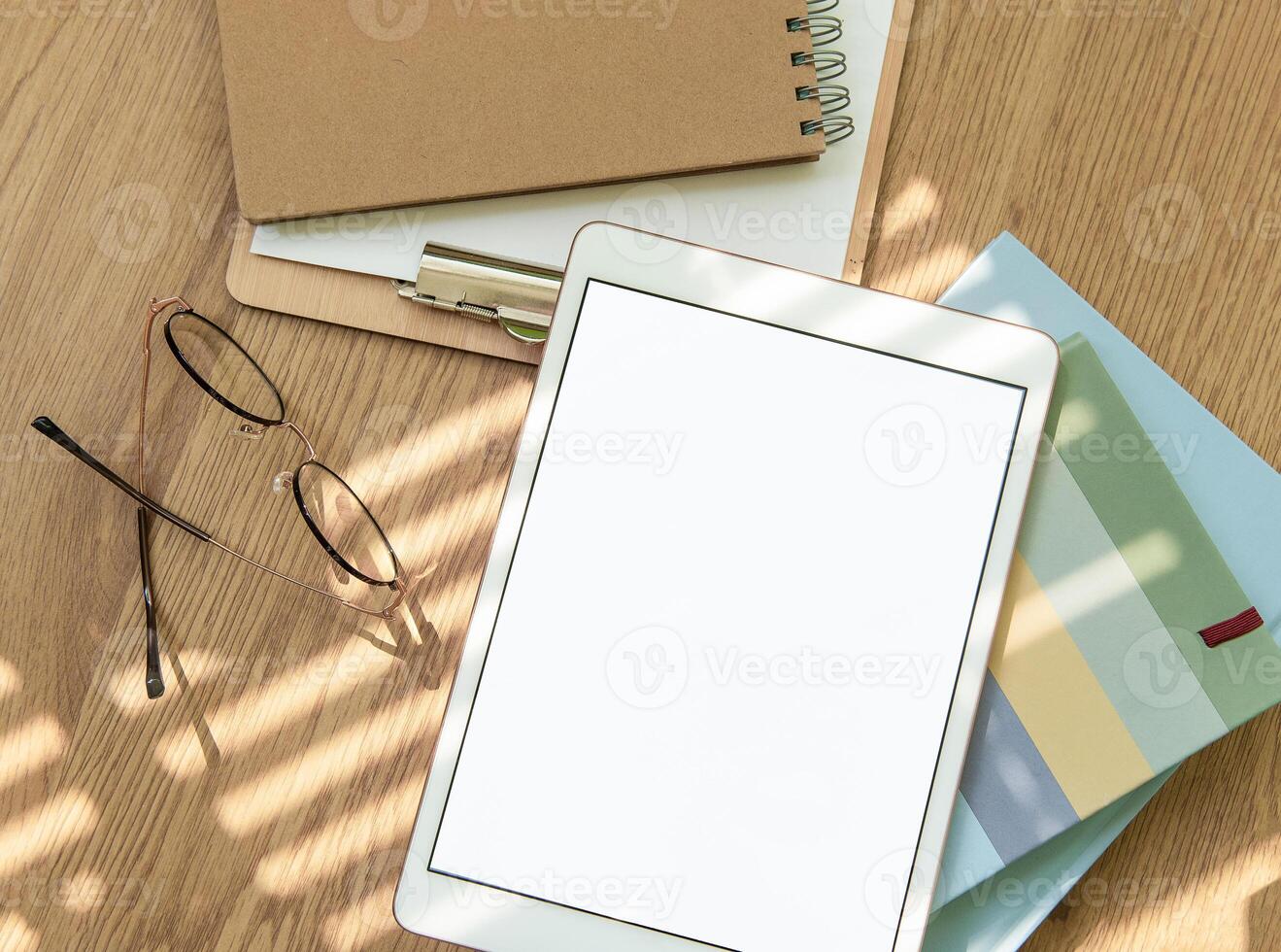 A workspace setup with a tablet, notebooks, glasses, and stationery on a wooden table illuminated by soft sunlight photo