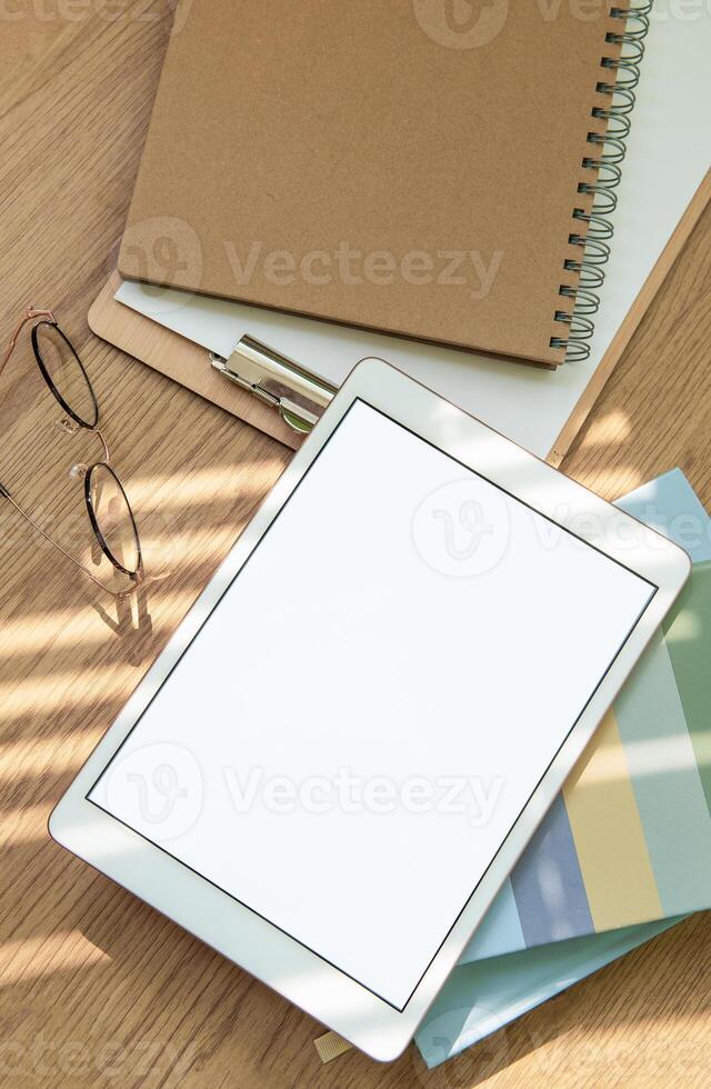 A workspace setup with a tablet, notebooks, glasses, and stationery on a wooden table illuminated by soft sunlight photo
