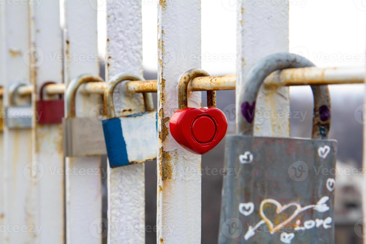 Close-up of various padlocks, including a red heart-shaped lock, attached to a rusty white metal railing symbolizing love, commitment, and memories on a bridge photo