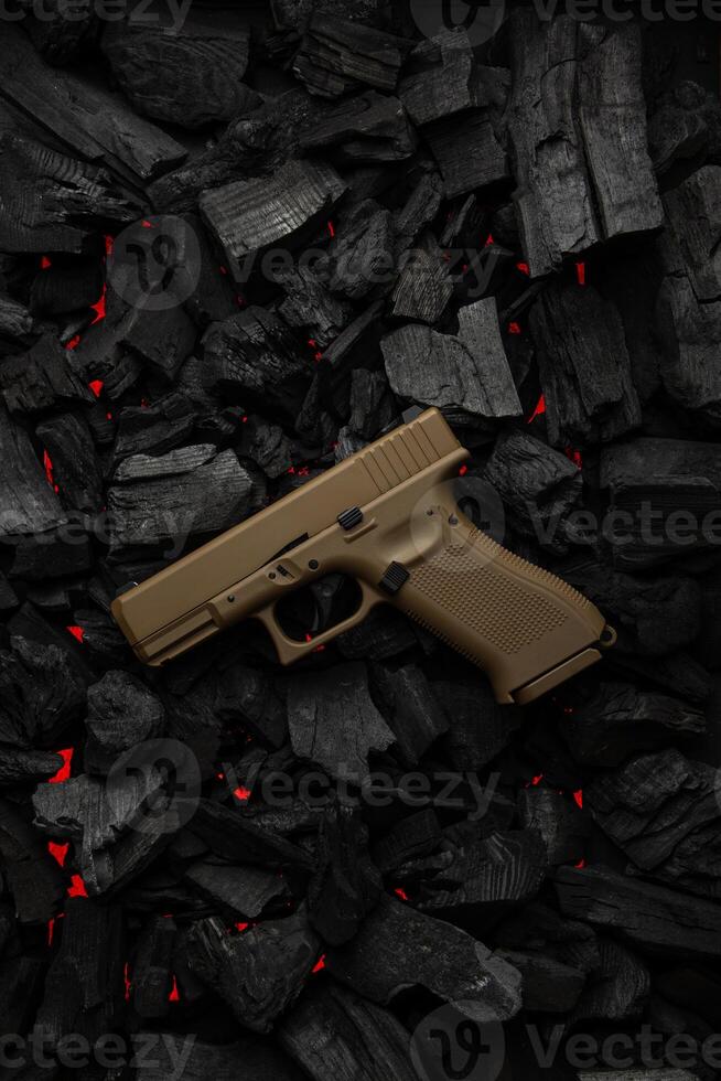 Close-up of a tan handgun placed on glowing charcoal, creating a contrast between the firearm's smooth surface and the rugged texture of the coal. Ideal for firearm and tactical themed content. photo
