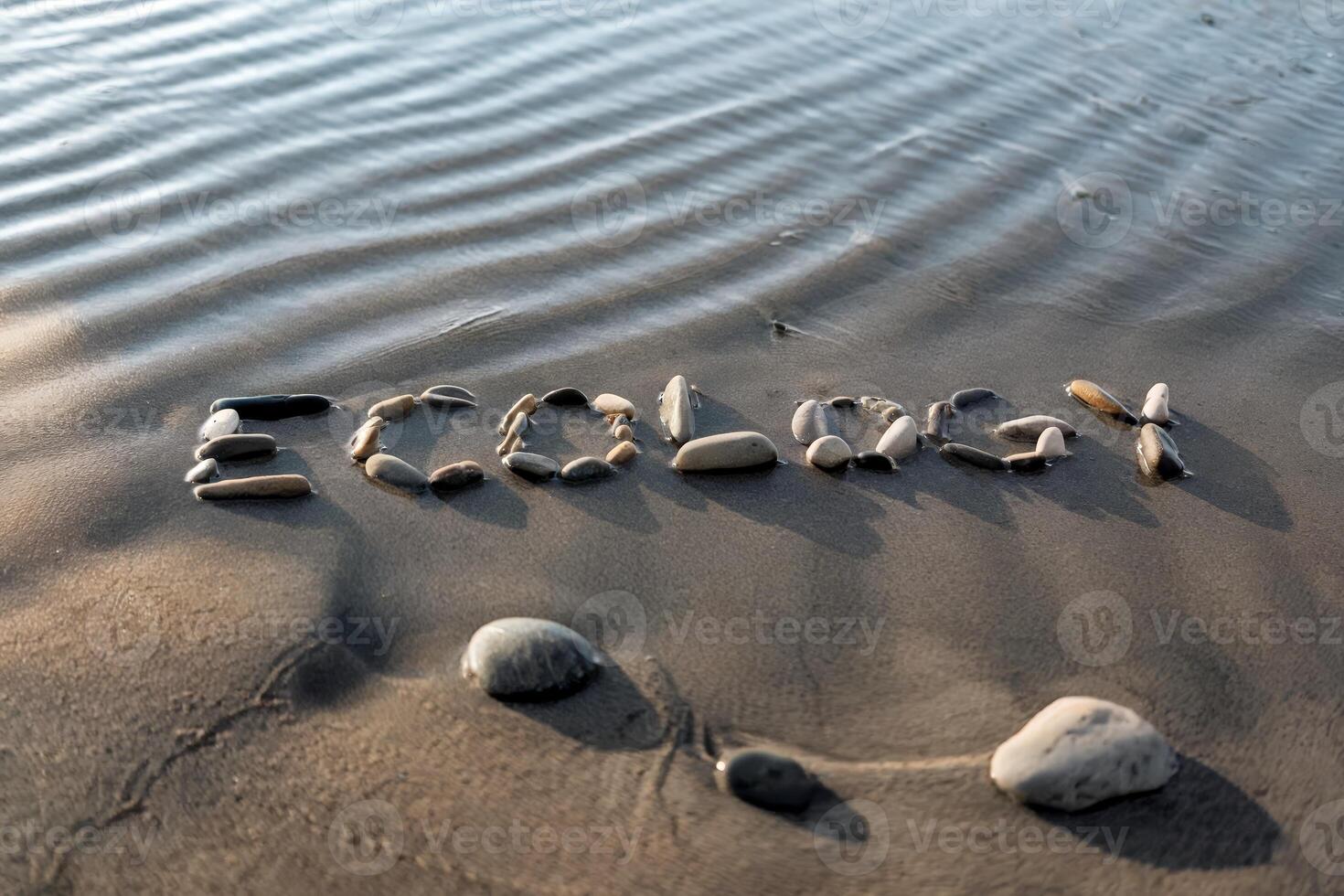 Stones arranged to spell 'Ecology' on a serene beach by rippling water. photo