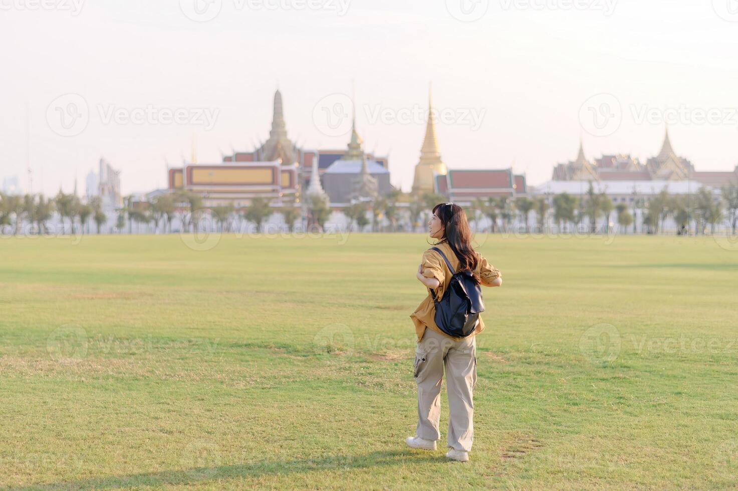 Traveler asian woman in her 30s, backpack slung over her shoulder, explores Wat Pra Kaew with childlike wonder. Sunlight dances on the golden rooftops, and a carefree smile photo