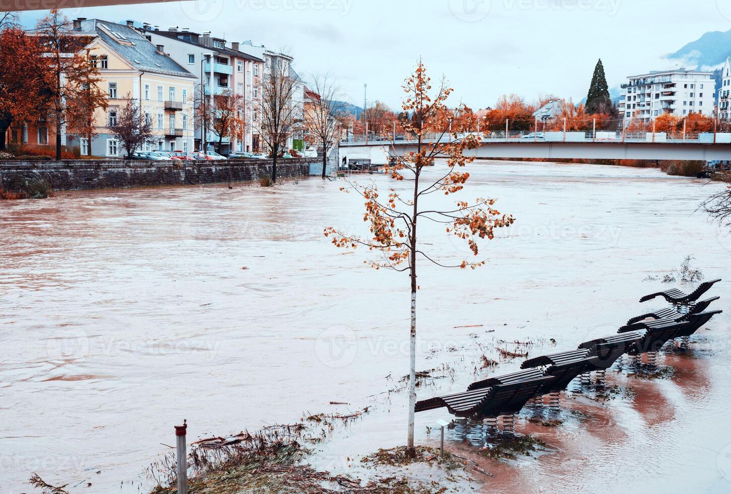 Flooding trees, street benches, near overflowing river, autumn cityscape in background, disaster concept photo