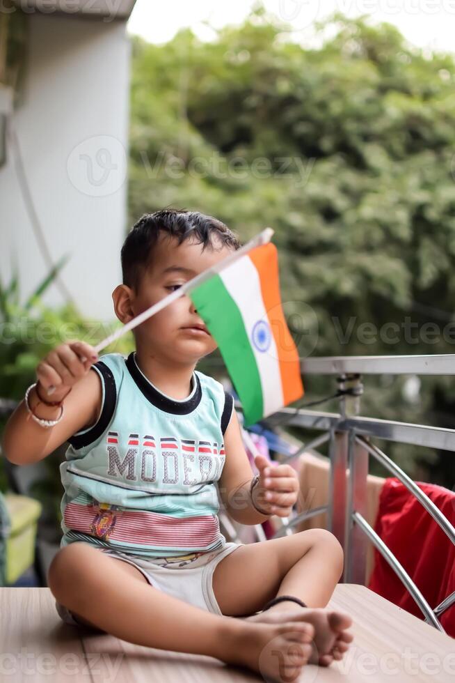 Cute Indian boy holding Indian National Flag in his hangs at home balcony. Bright portrait of happy child sitting on the table with national flag photo