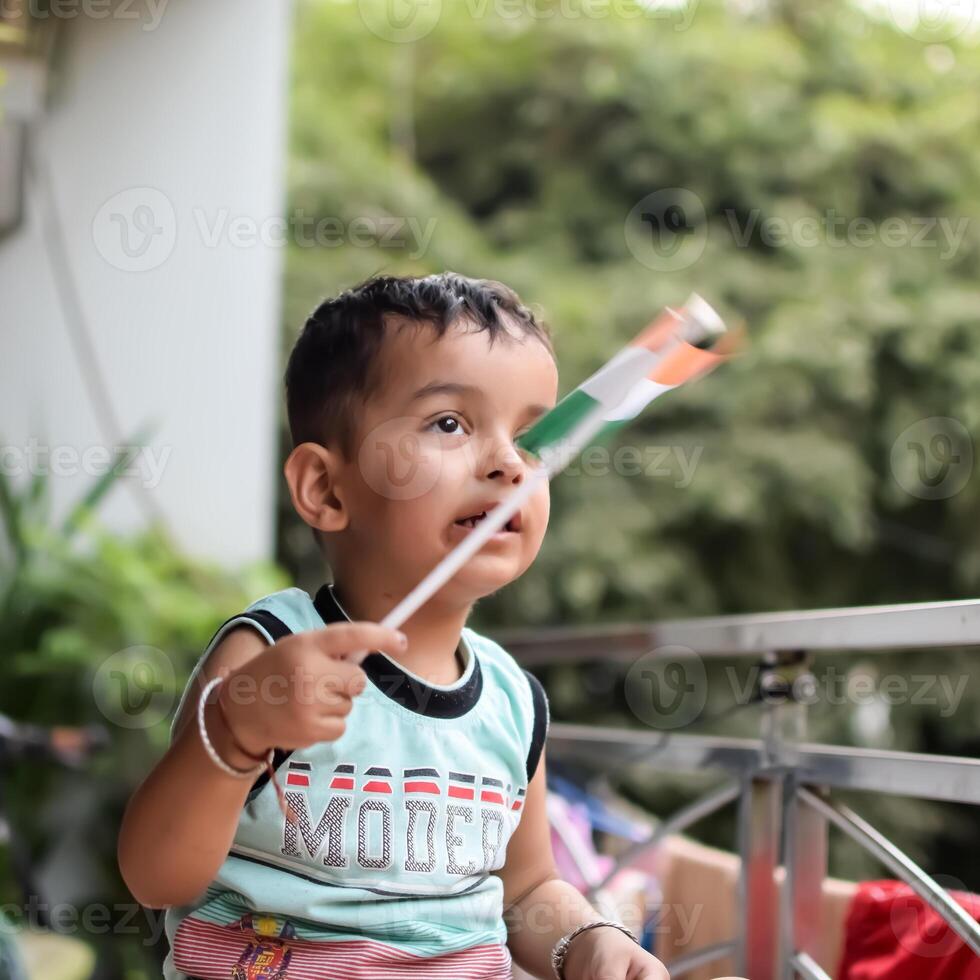 Cute Indian boy holding Indian National Flag in his hangs at home balcony. Bright portrait of happy child sitting on the table with national flag photo