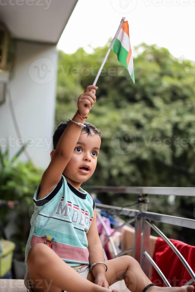 Cute Indian boy holding Indian National Flag in his hangs at home balcony. Bright portrait of happy child sitting on the table with national flag photo
