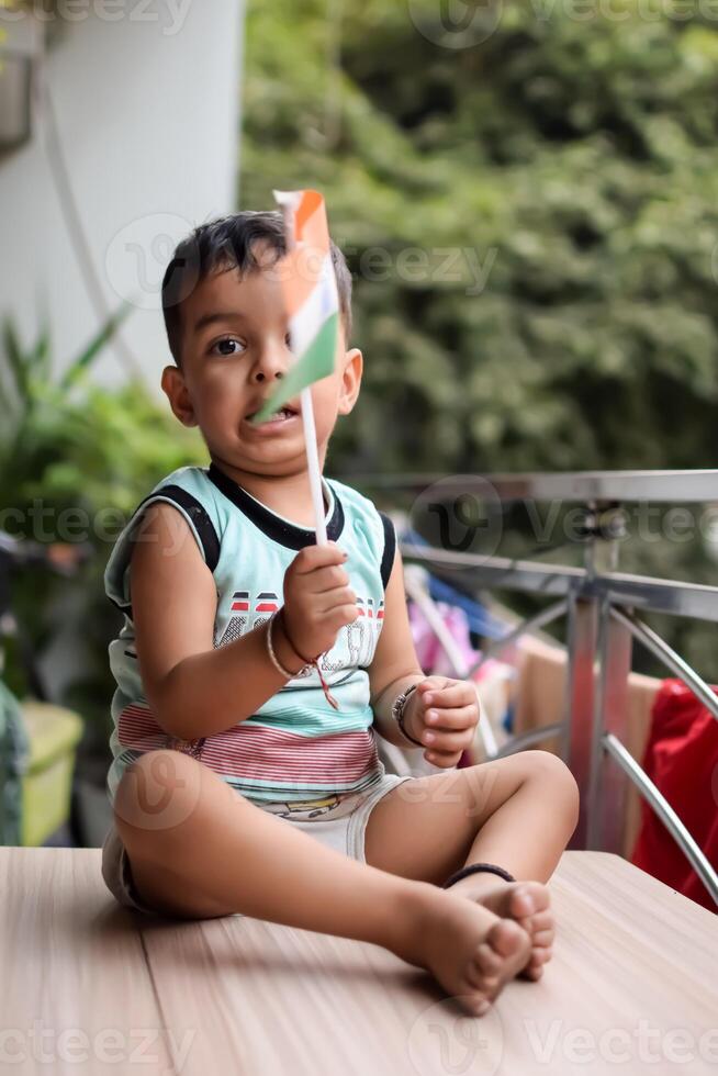Cute Indian boy holding Indian National Flag in his hangs at home balcony. Bright portrait of happy child sitting on the table with national flag photo
