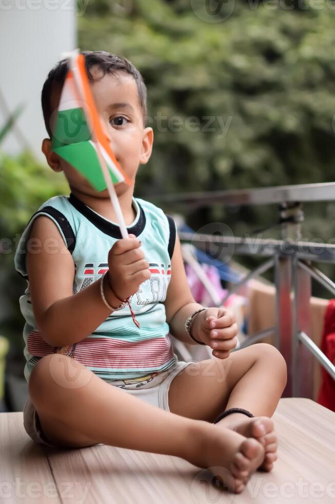 Cute Indian boy holding Indian National Flag in his hangs at home balcony. Bright portrait of happy child sitting on the table with national flag photo
