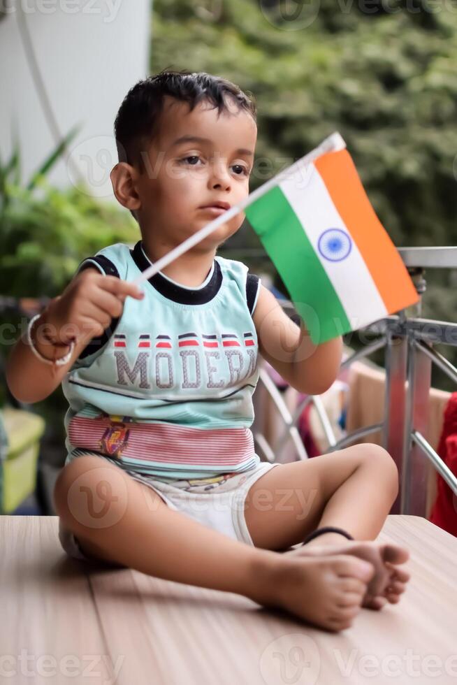Cute Indian boy holding Indian National Flag in his hangs at home balcony. Bright portrait of happy child sitting on the table with national flag photo