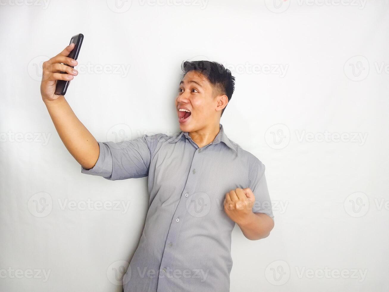Young Asian man showing wow face expression when looking to his mobile phone wearing grey shirt isolated on white background photo